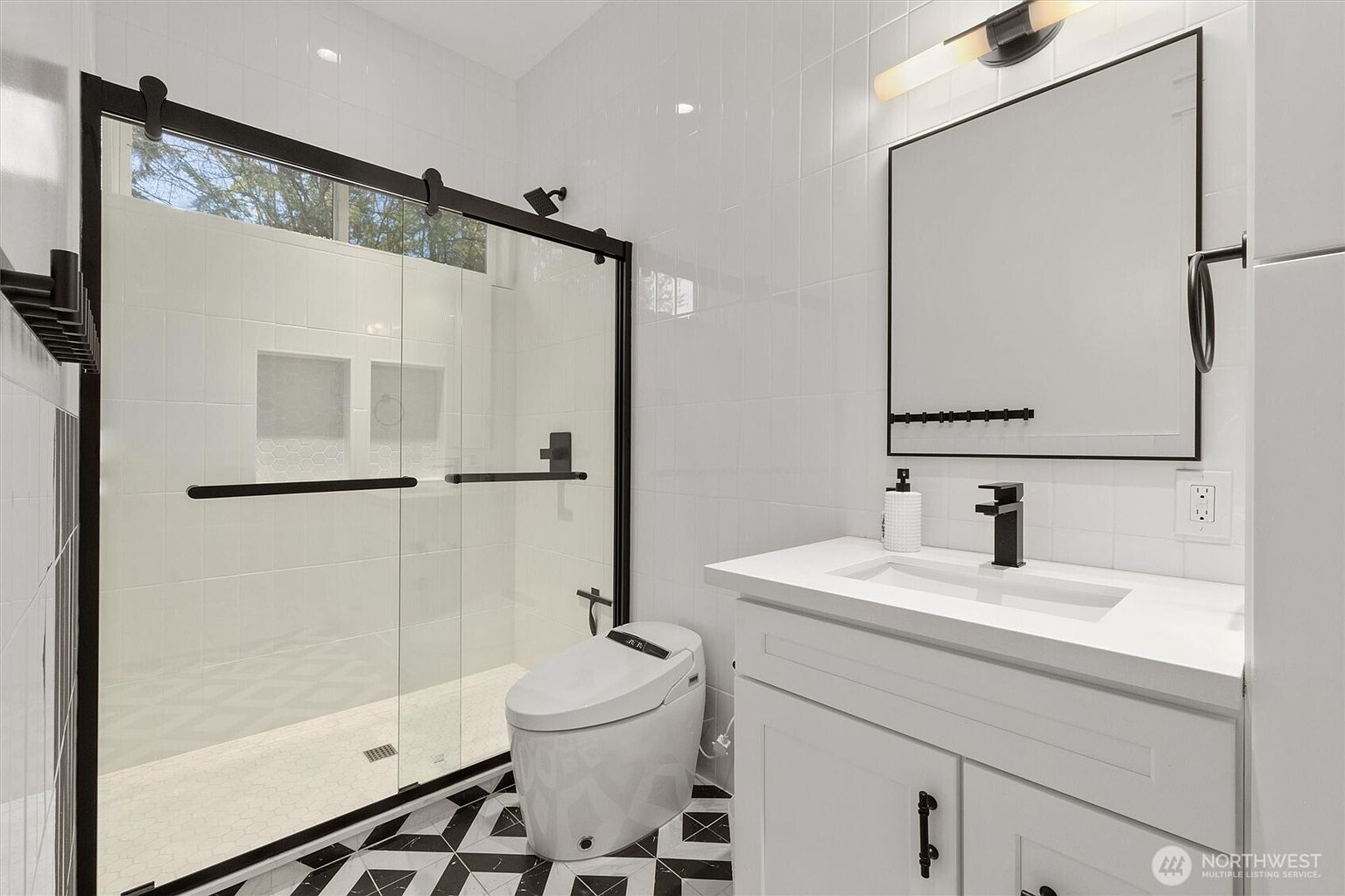 This is a modern primary bathroom featuring a glass-enclosed shower with black trim, a white toilet, and a white vanity with a black faucet. The floor is tiled in a black and white geometric pattern, and the walls are white tiled. The overall impression is clean, stylish, and well-maintained.