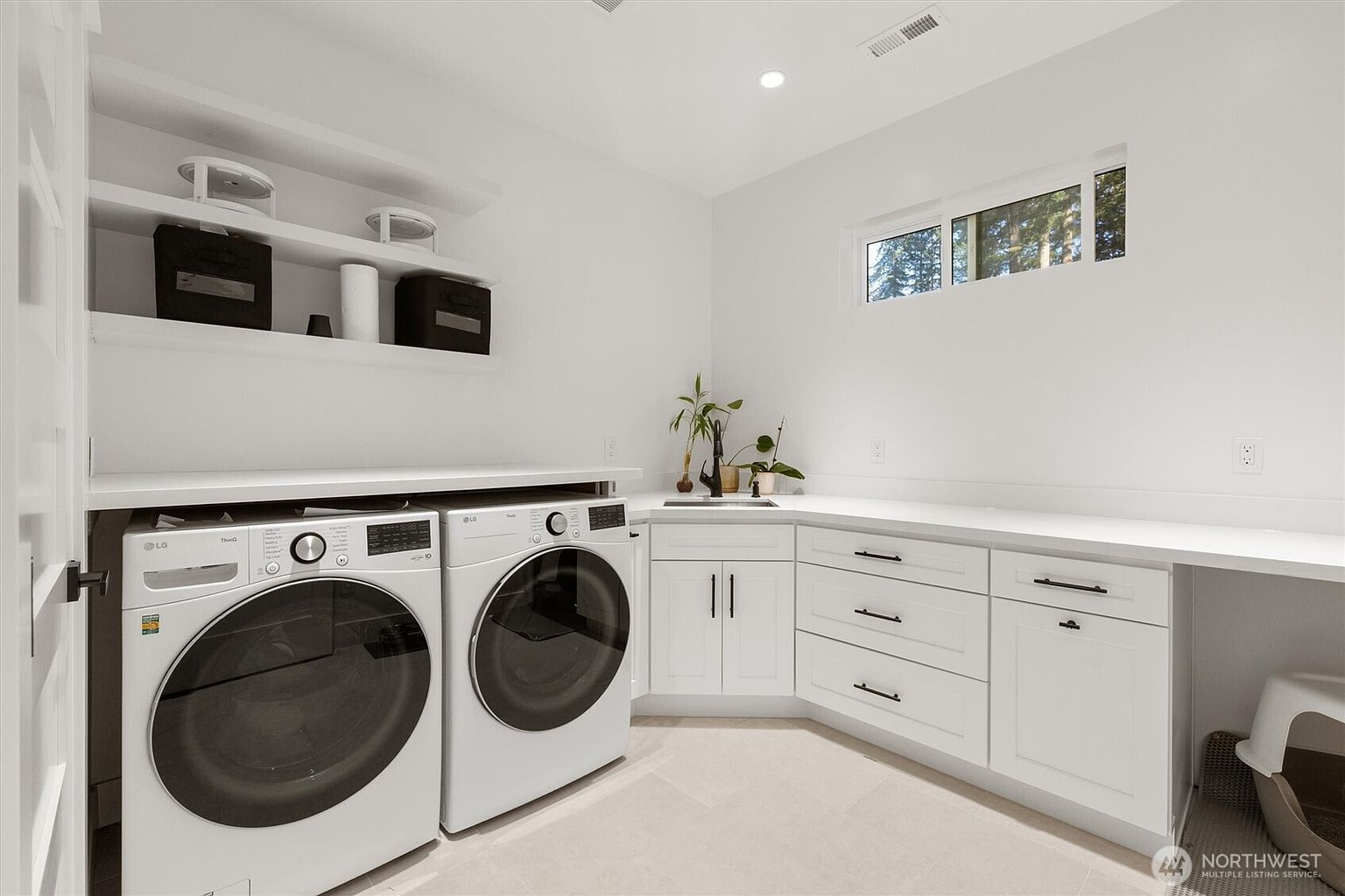 This is a bright and modern laundry room featuring white walls, cabinetry, and countertops. The room includes a front-loading washer and dryer set, a sink with a black faucet, and open shelving for storage. A window provides natural light, and the overall impression is clean and functional.