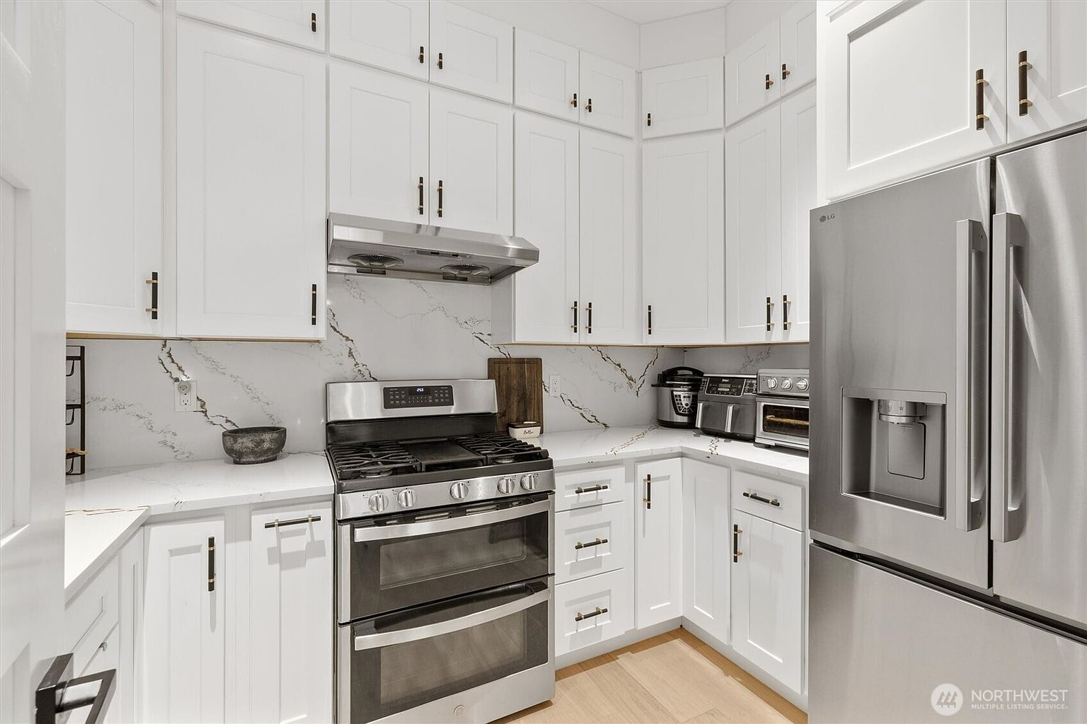 This is a well-lit kitchen featuring white cabinetry with bronze hardware, stainless steel appliances, and light wood flooring. The countertops and backsplash are a matching white marble with grey veining. The kitchen has a modern and clean aesthetic, making it an inviting space for cooking and entertaining.