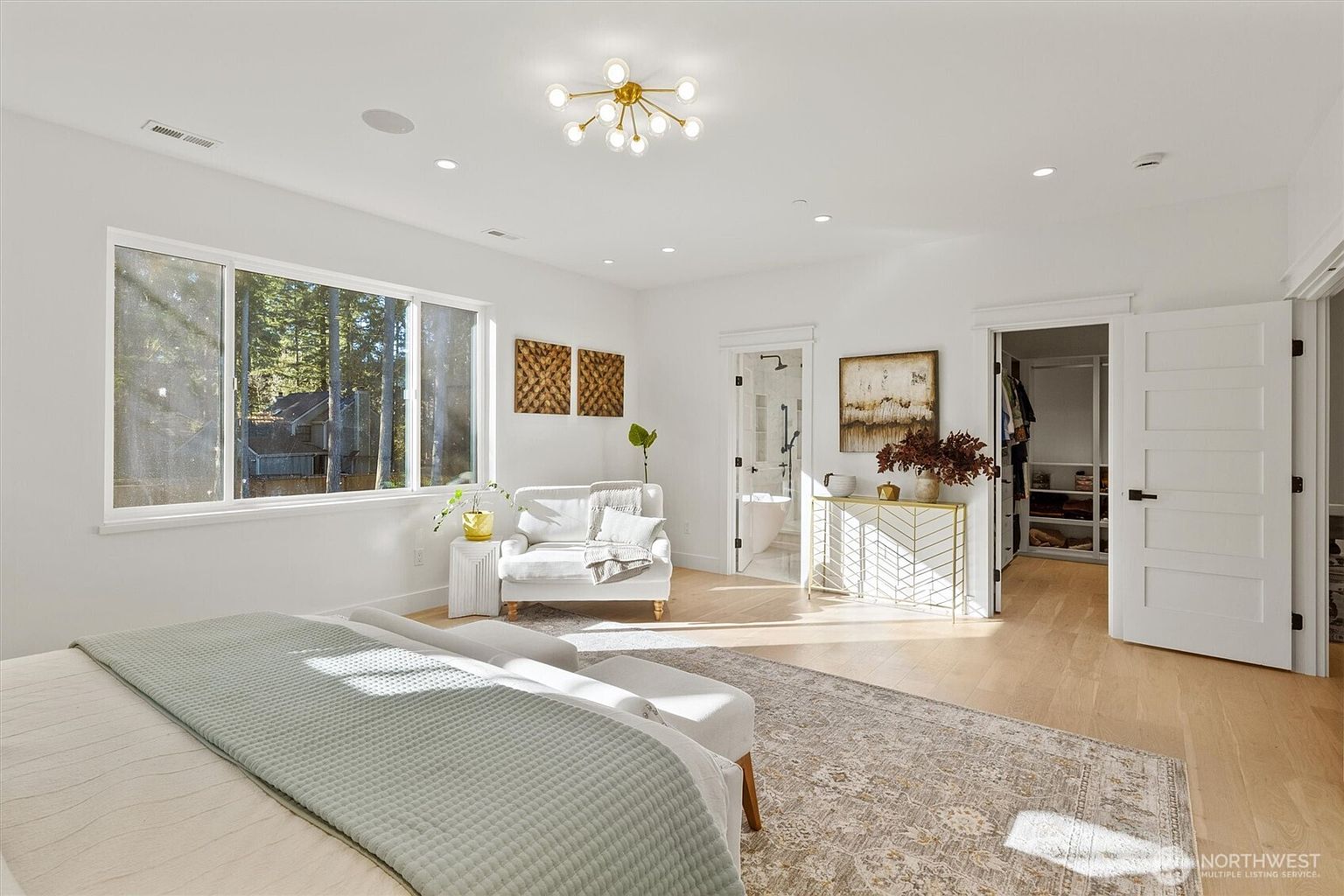 This is a bright and spacious primary bedroom featuring a large window with natural light, a seating area with a white loveseat, and a neutral-toned rug. The room has a clean, modern aesthetic with white walls and light wood flooring, complemented by stylish light fixtures and artwork. The open doorway leads to a walk-in closet and a bathroom, enhancing the suite-like feel.