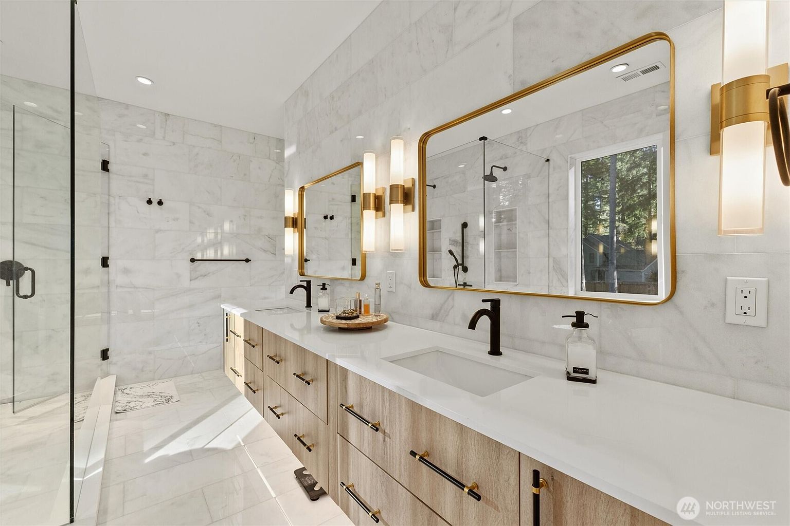 This is a luxurious primary bathroom featuring a double vanity with a light wood finish and black hardware, topped with a white countertop and dual sinks. Gold-framed mirrors and sconces add a touch of elegance, while the marble-tiled walls and floors create a spa-like atmosphere. A glass-enclosed shower is visible on the left, enhancing the modern and clean aesthetic.