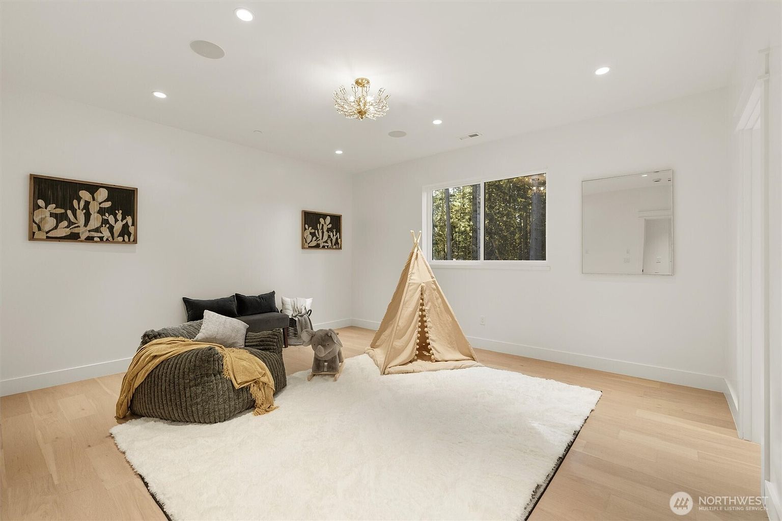 This is a bright and airy children's bedroom featuring white walls and light wood flooring. The room is decorated with a teepee, a plush rug, and playful artwork, creating a cozy and inviting atmosphere. A comfortable seating area with a beanbag chair and a small sofa adds to the room's functionality and charm.