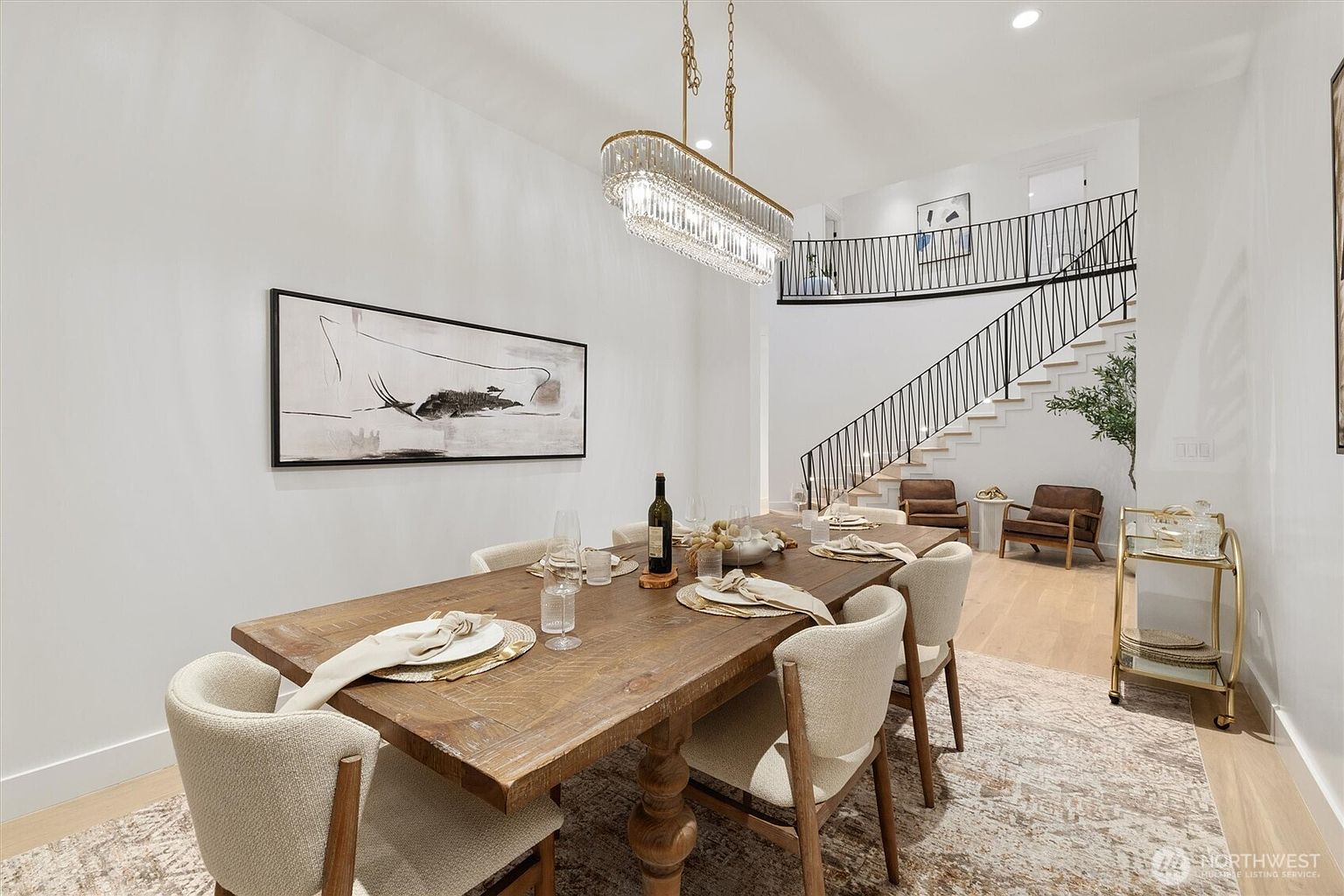This interior shot showcases a dining room with a large wooden table set for a meal, surrounded by upholstered chairs. A modern chandelier hangs above the table, and an abstract painting adorns the wall. The room features a staircase in the background, adding architectural interest and suggesting a spacious, open floor plan.