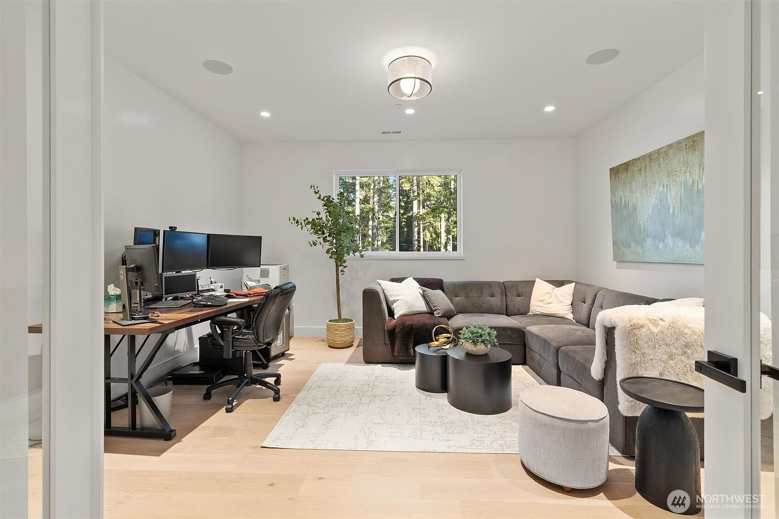 This interior shot showcases a well-lit office/study area featuring a modern aesthetic. A large desk with multiple monitors and an ergonomic chair are positioned to the left, while a comfortable gray sectional sofa anchors the right side of the room. Natural light streams in through a window, complementing the neutral color palette and creating a serene and productive atmosphere.