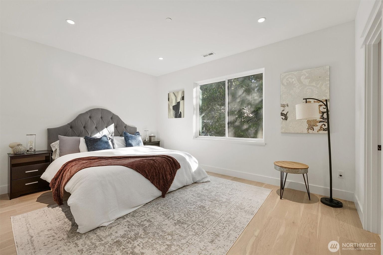 This is a bright and airy primary bedroom featuring a gray upholstered headboard, a white comforter with a brown throw, and blue decorative pillows. The room is well-lit with recessed lighting and natural light from a window, complemented by a neutral-toned rug and light wood flooring. Artwork adorns the walls, adding a touch of sophistication to the space.
