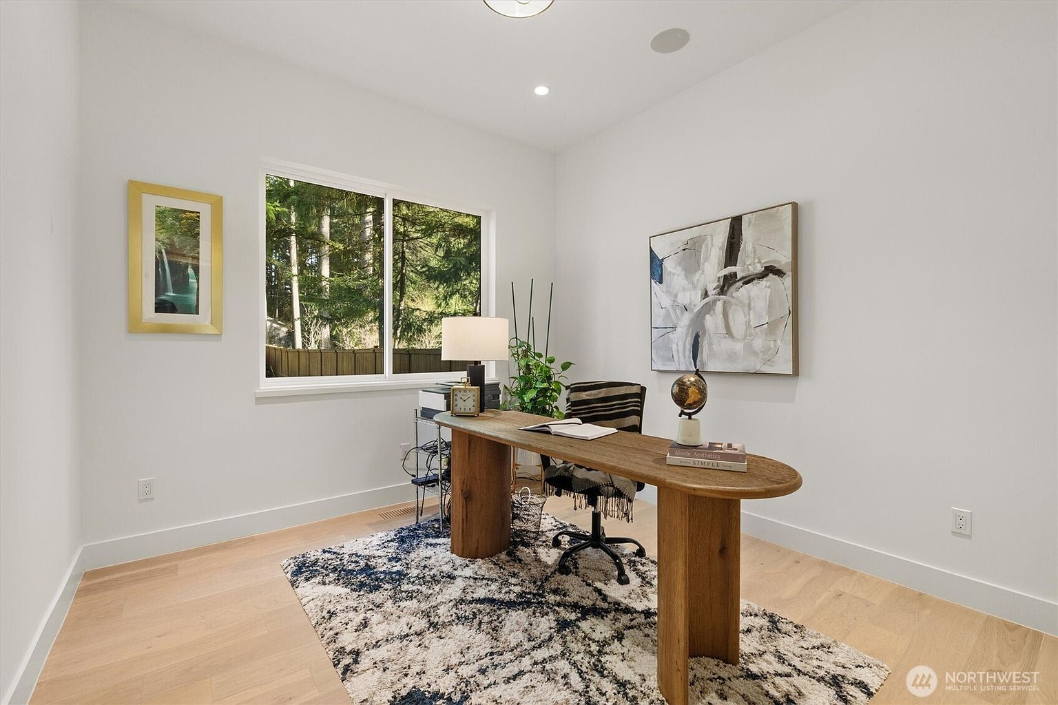 This is an interior shot of a bright and modern office space. The room features a wooden desk with a unique design, a comfortable office chair, and a large window providing natural light and a view of the outdoors. Artwork adorns the walls, and a patterned rug adds texture to the light wood flooring, creating a stylish and functional workspace.