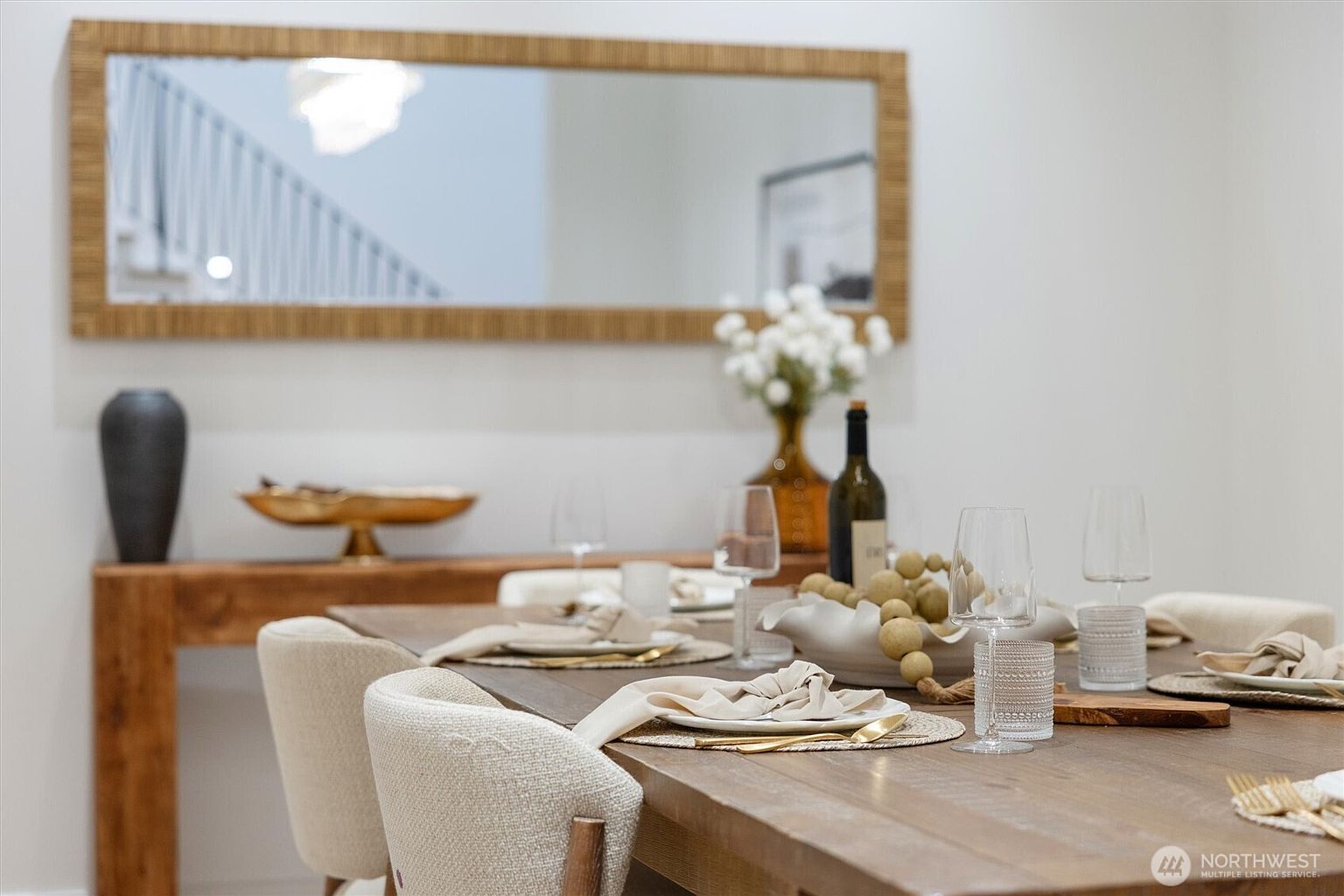 This is an interior shot of a dining room featuring a wooden table set with elegant place settings, including plates, napkins, glassware, and gold cutlery. A console table behind the dining table is decorated with a vase, a decorative bowl, and a framed mirror reflecting a staircase. The room has a neutral color palette, creating a sophisticated and inviting atmosphere.