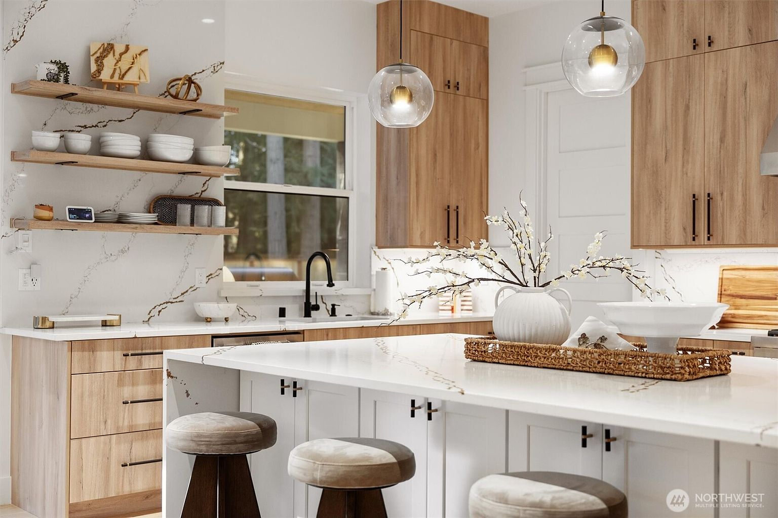 This is a bright and modern kitchen featuring light wood cabinetry, white countertops with subtle veining, and a central island with seating. Pendant lights hang above the island, and open shelving displays decorative items and dishware. The kitchen has a clean and contemporary design, creating a welcoming and functional space.