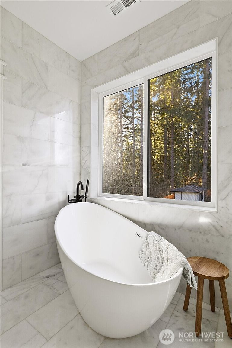 This is a bright and airy primary bathroom featuring a freestanding white bathtub with a black faucet. The walls and floor are covered in marble tiles, and a large window provides natural light and a view of the trees outside. A small wooden stool sits next to the tub, adding a touch of warmth to the space.
