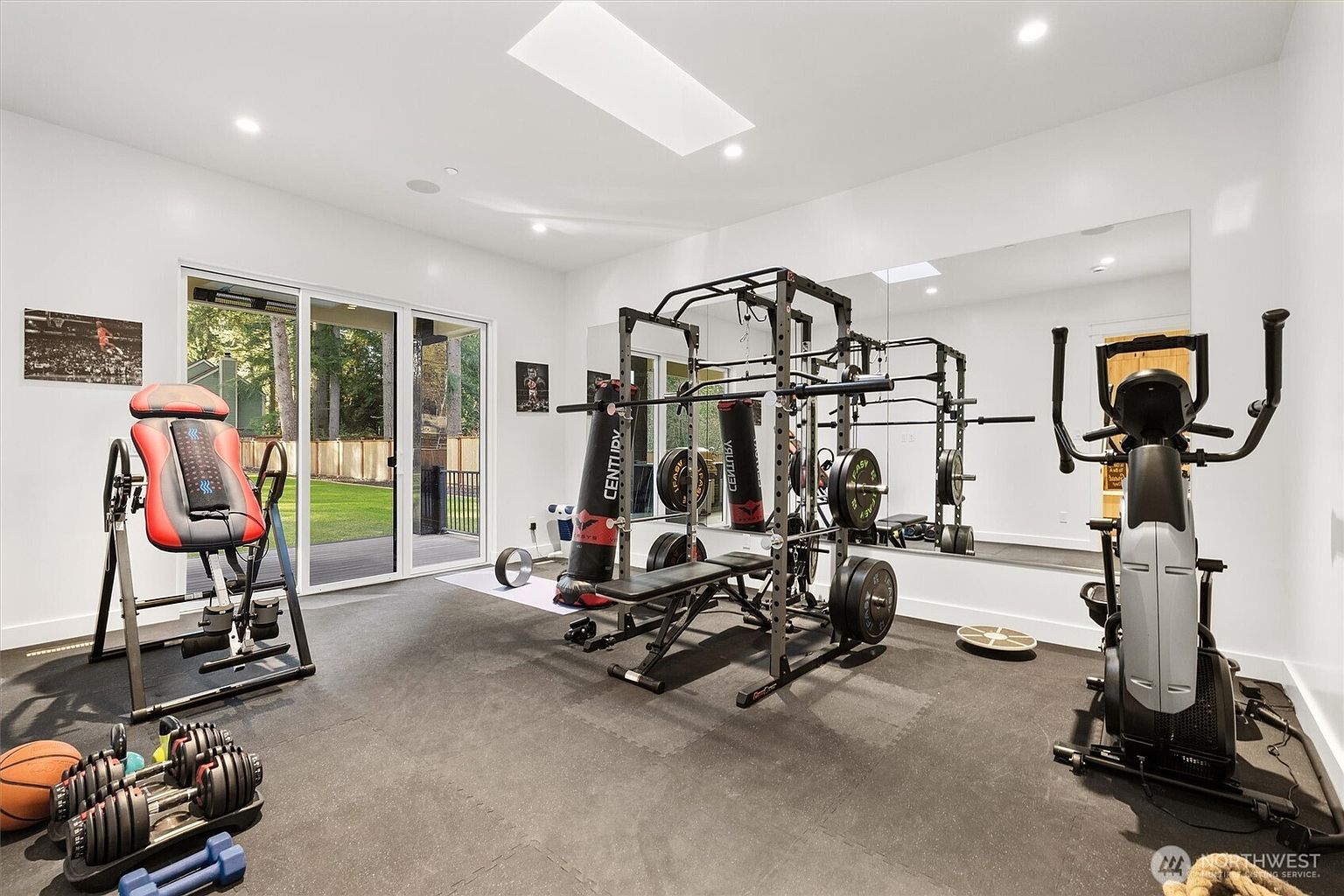 This is an interior shot of a well-equipped home gym. The room features a variety of exercise equipment, including a weight rack, punching bag, exercise bike, and inversion table. The flooring is a dark rubber mat, and the walls are painted white, creating a bright and functional workout space.