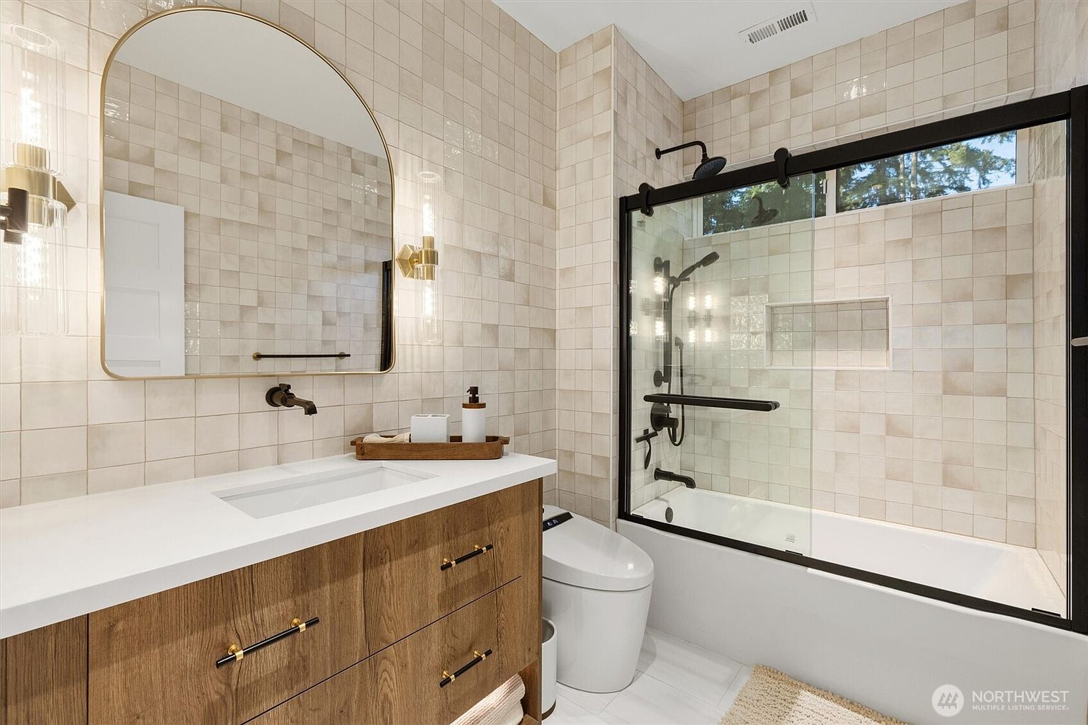 This is a well-lit bathroom featuring a modern design with neutral tones. The vanity has a wood-grain cabinet with a white countertop and a rectangular sink. A large, arched mirror hangs above the sink, flanked by sconce lighting, and a glass-enclosed shower/tub combination is visible to the right, creating a clean and inviting space.