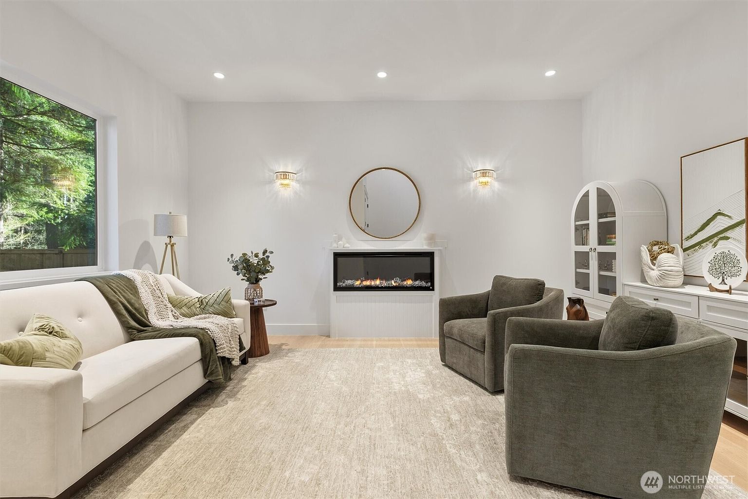 This is a bright and airy living room featuring a modern fireplace with a round mirror above it, flanked by sconces. The room is furnished with a light-colored sofa, two armchairs, and a neutral-toned rug, creating a comfortable and inviting space. A large window provides natural light and a view of the outdoors.