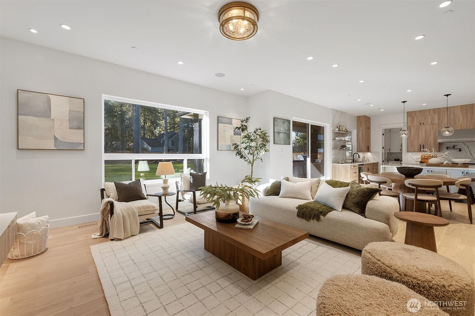 This is an interior shot of a modern living room featuring a neutral color palette. The room is well-lit with recessed lighting and natural light from a large window overlooking a green landscape. Key features include a comfortable sofa with decorative pillows, a wooden coffee table with decorative items, and a patterned area rug, creating a cozy and inviting atmosphere.