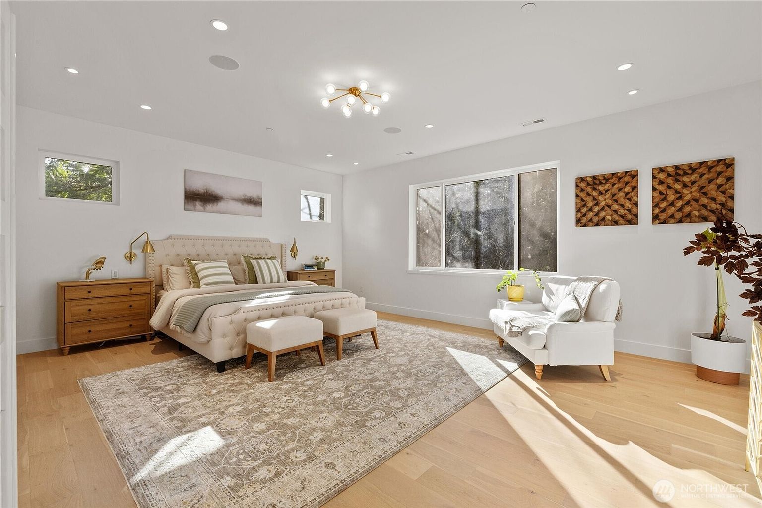 This is a bright and spacious primary bedroom featuring a large bed with a tufted headboard, complemented by matching ottomans and wooden nightstands. The room is decorated with neutral tones and accented by a patterned rug, creating a serene and inviting atmosphere. Natural light floods the space through a large window, highlighting the hardwood floors and modern light fixture.