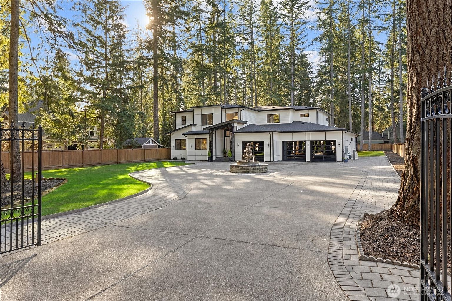 This is a front exterior view of a modern two-story home with a black and white color scheme. The property features a circular driveway with a fountain in the center, a well-manicured lawn, and a gated entrance. Tall trees surround the house, providing privacy and a sense of seclusion.