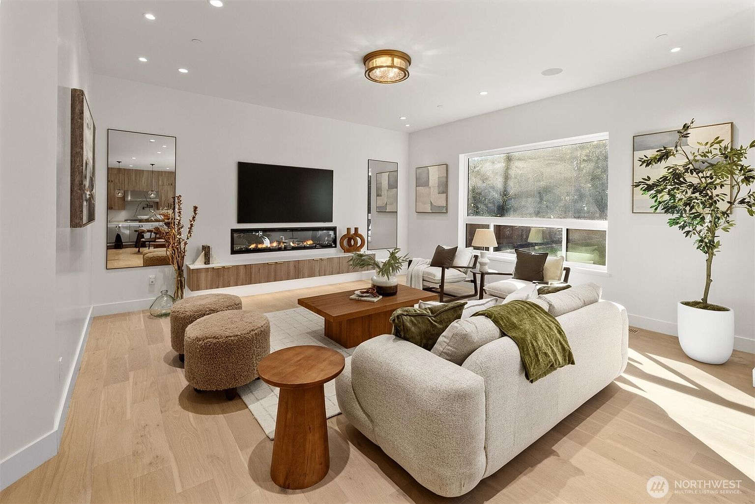 This is a bright and modern living room featuring a large flat-screen TV above a linear fireplace, complemented by stylish wooden cabinetry. The room is furnished with a comfortable-looking sofa, accent chairs, and wooden coffee table, all set on a light-colored hardwood floor. Natural light floods the space through a large window, enhancing the room's inviting and contemporary atmosphere.