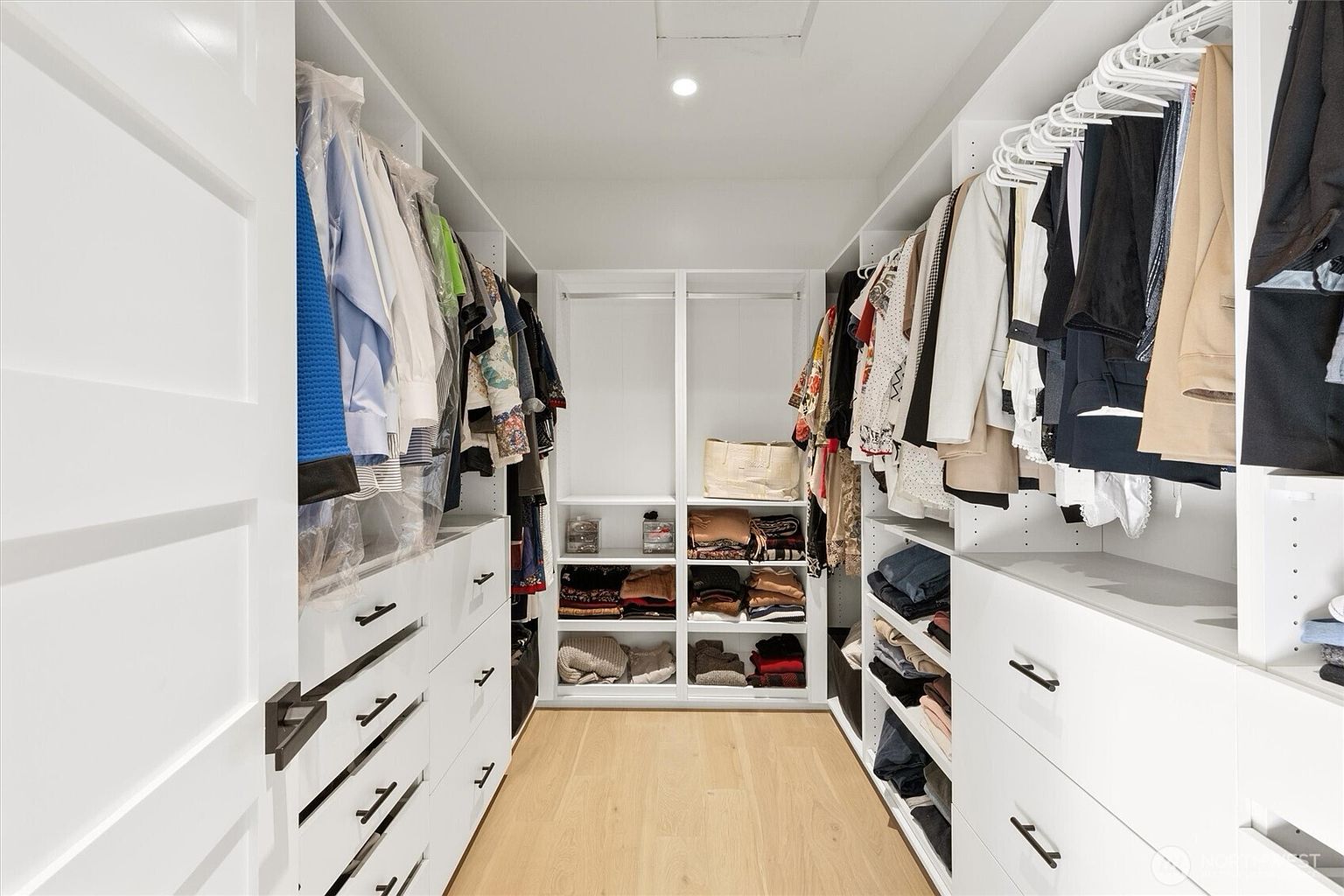 This is a well-organized walk-in closet featuring white cabinetry and drawers with dark hardware. Clothes are neatly hung on hangers, and folded items are arranged on shelves. The closet has a light wood floor and is brightly lit, creating a clean and functional space.