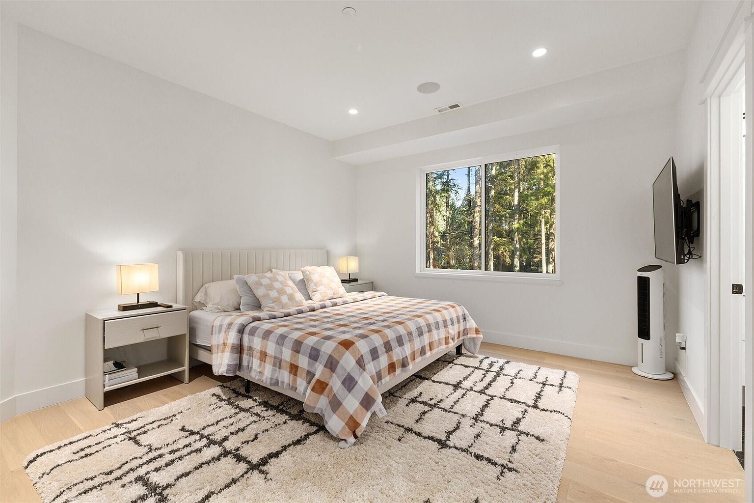 This is a bright and airy bedroom featuring a queen-sized bed with a plaid comforter and decorative pillows. The room has light wood flooring, white walls, and a large window that provides natural light and a view of the trees outside. A modern nightstand with a lamp sits on either side of the bed, and a patterned rug adds texture to the space.
