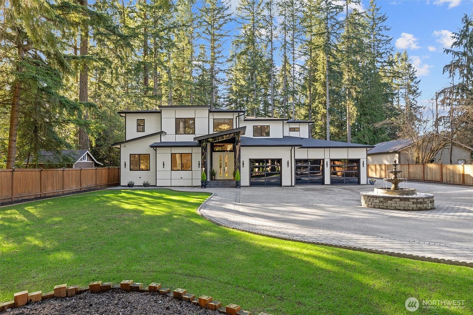 This is a front exterior view of a modern two-story home with a well-manicured lawn and mature trees in the background. The house features a white facade with black trim, multiple windows, and a three-car garage with glass doors. A circular fountain sits in the driveway, adding a touch of elegance to the property.