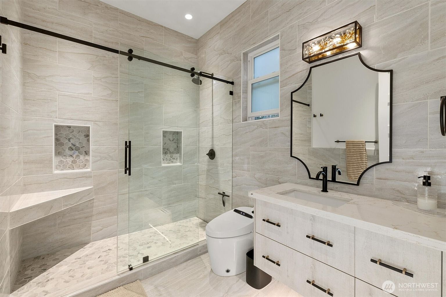 This is a well-lit primary bathroom featuring a modern design with light-colored, horizontally laid tiles on the walls and floor. A glass-enclosed shower with a built-in bench and niche is on the left, while a contemporary vanity with a white countertop, dark hardware, and a uniquely shaped mirror is on the right. The bathroom also includes a modern toilet and a decorative light fixture above the mirror, creating a spa-like atmosphere.