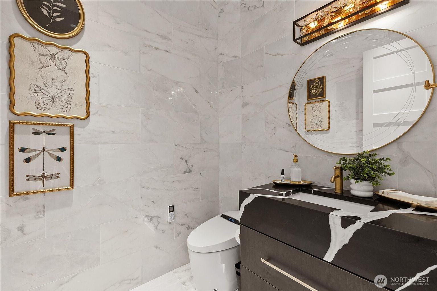 This is a stylish guest bathroom featuring marble-like tile walls and flooring, creating a luxurious and clean aesthetic. A modern vanity with a dark finish and a white countertop with striking marble veining is complemented by a round mirror and decorative lighting. The toilet is a sleek, modern design, and framed artwork adds a touch of elegance to the space.