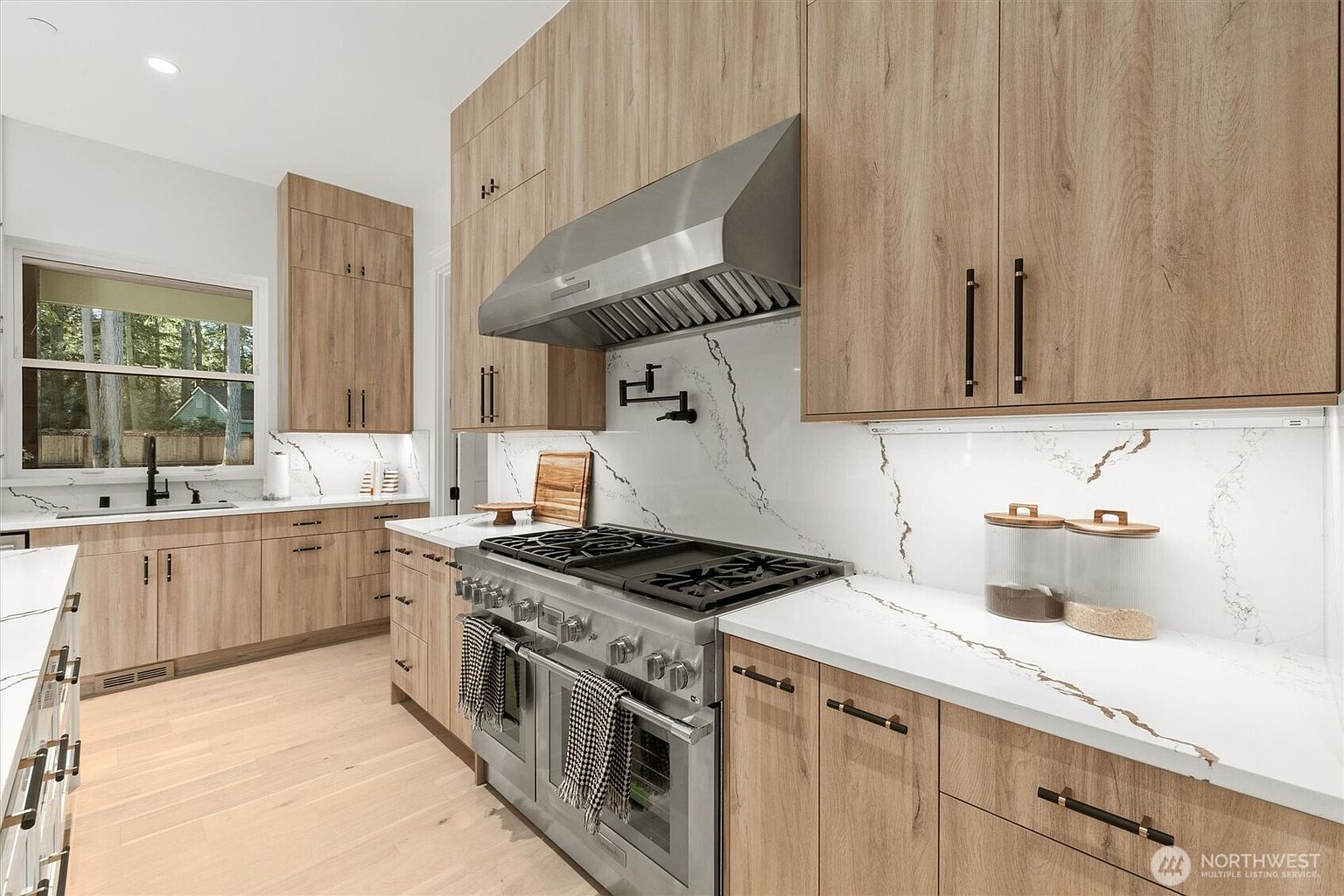 This is a well-lit kitchen featuring light wood cabinetry with sleek, dark hardware. The countertops are white with marble veining, complementing the backsplash. A stainless steel range hood and appliances add a modern touch, while the light wood flooring contributes to a bright and airy atmosphere.