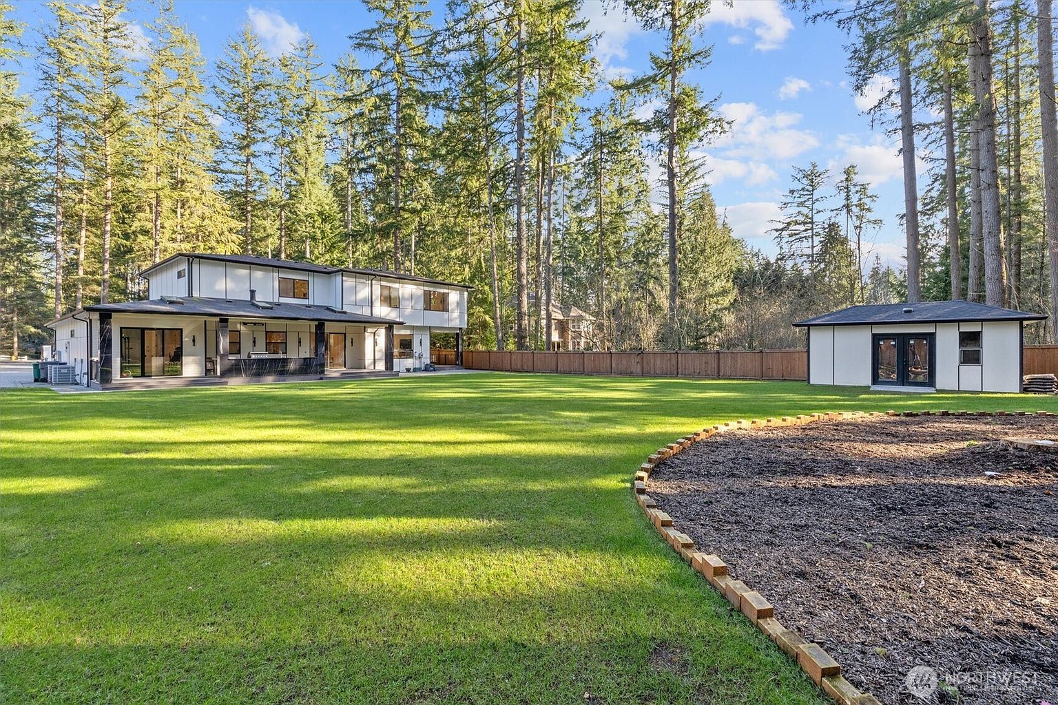 This image showcases the expansive backyard of a modern home, featuring a lush green lawn that stretches towards a two-story house with a dark-trimmed exterior. A separate outbuilding is visible on the right, and a wooden fence encloses the property, providing privacy. Tall trees surround the yard, creating a serene and natural setting.