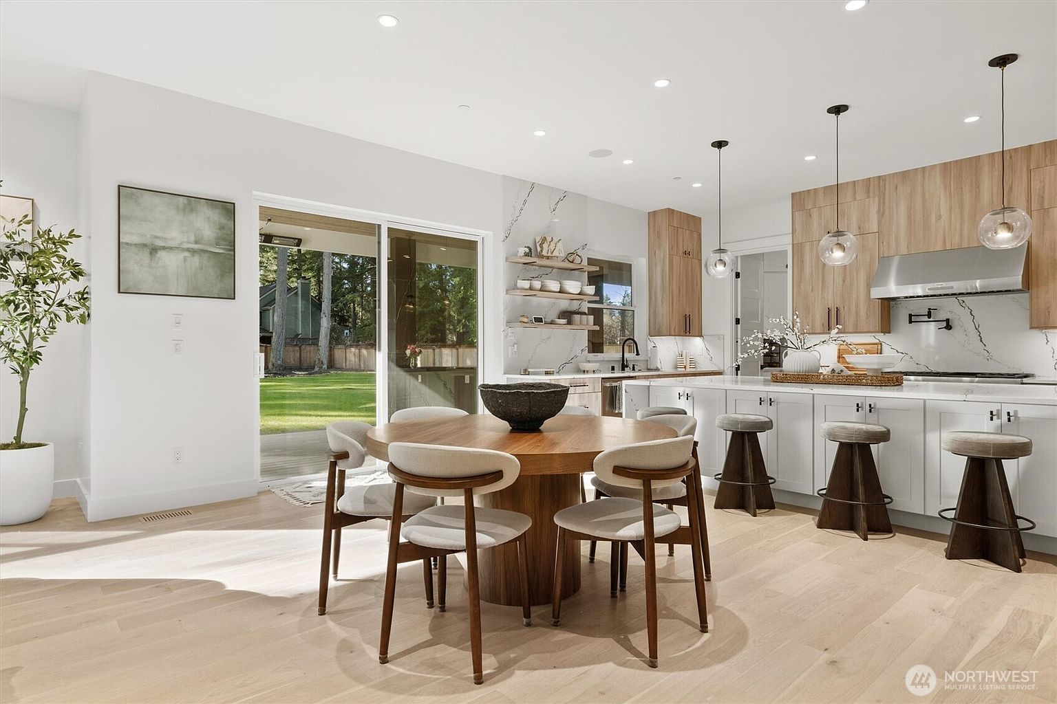 This interior shot showcases a modern kitchen and dining area with an open-concept design. The kitchen features white and wood-tone cabinetry, marble countertops and backsplash, and stainless steel appliances, complemented by pendant lighting. A round wooden dining table with upholstered chairs sits adjacent to the kitchen, creating a cohesive and inviting space.