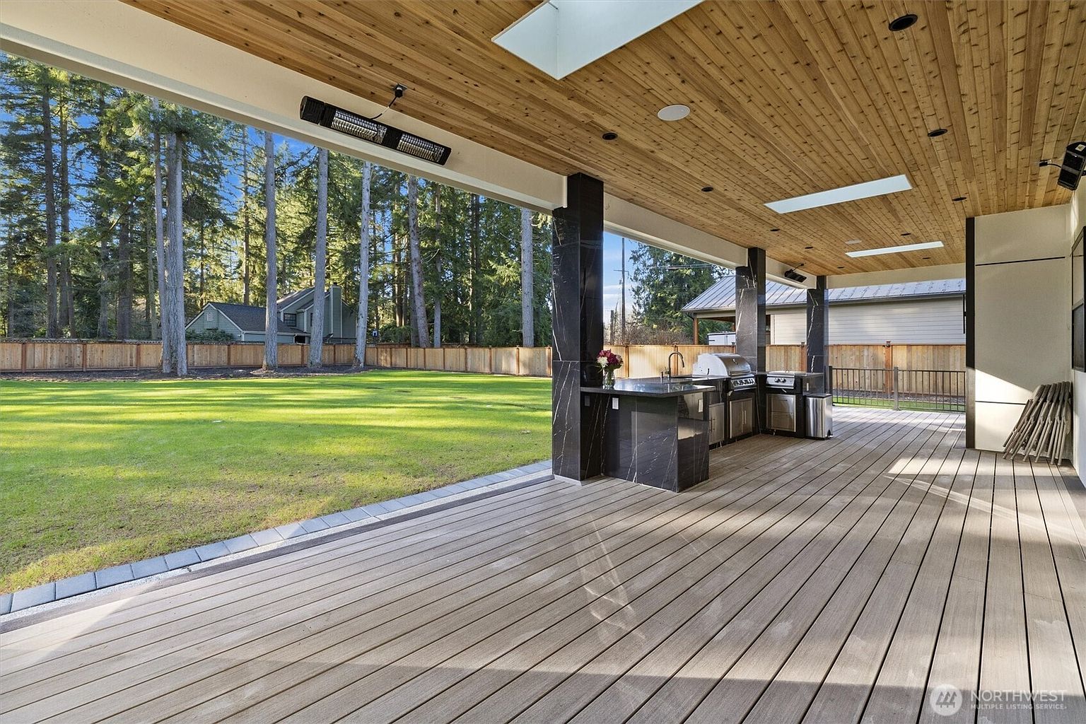 This image showcases a luxurious outdoor patio and deck area, complete with a built-in kitchen featuring granite countertops and stainless steel appliances. The deck is constructed with light-colored wood and offers a seamless transition to a well-manicured lawn and wooded backyard. The space is covered by a wooden ceiling with skylights and a mounted heater, creating an inviting and functional outdoor living area.