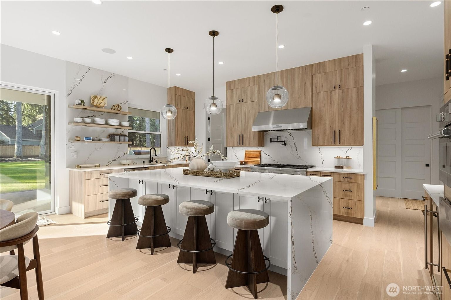 This is a bright and modern kitchen featuring light wood cabinetry and a large white island with seating. The countertops are white with subtle veining, and pendant lights hang above the island. The kitchen also features stainless steel appliances and open shelving, creating a clean and inviting space.