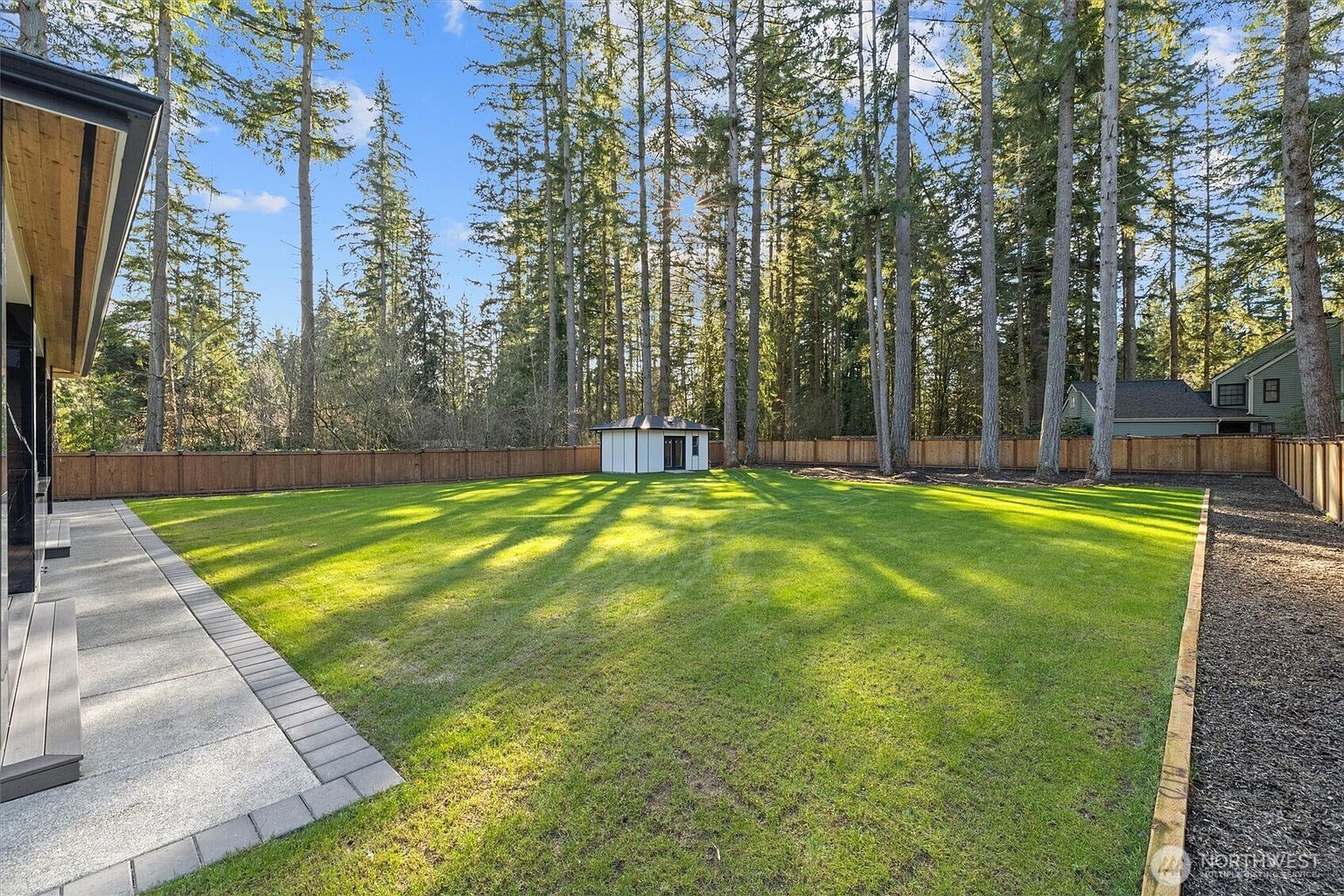 This image showcases a spacious and well-maintained backyard. The lush green lawn is surrounded by a wooden fence and tall trees, providing privacy and a natural setting. A small white shed is visible in the distance, adding a touch of charm to the landscape. The perspective is a wide shot, emphasizing the size and openness of the yard.