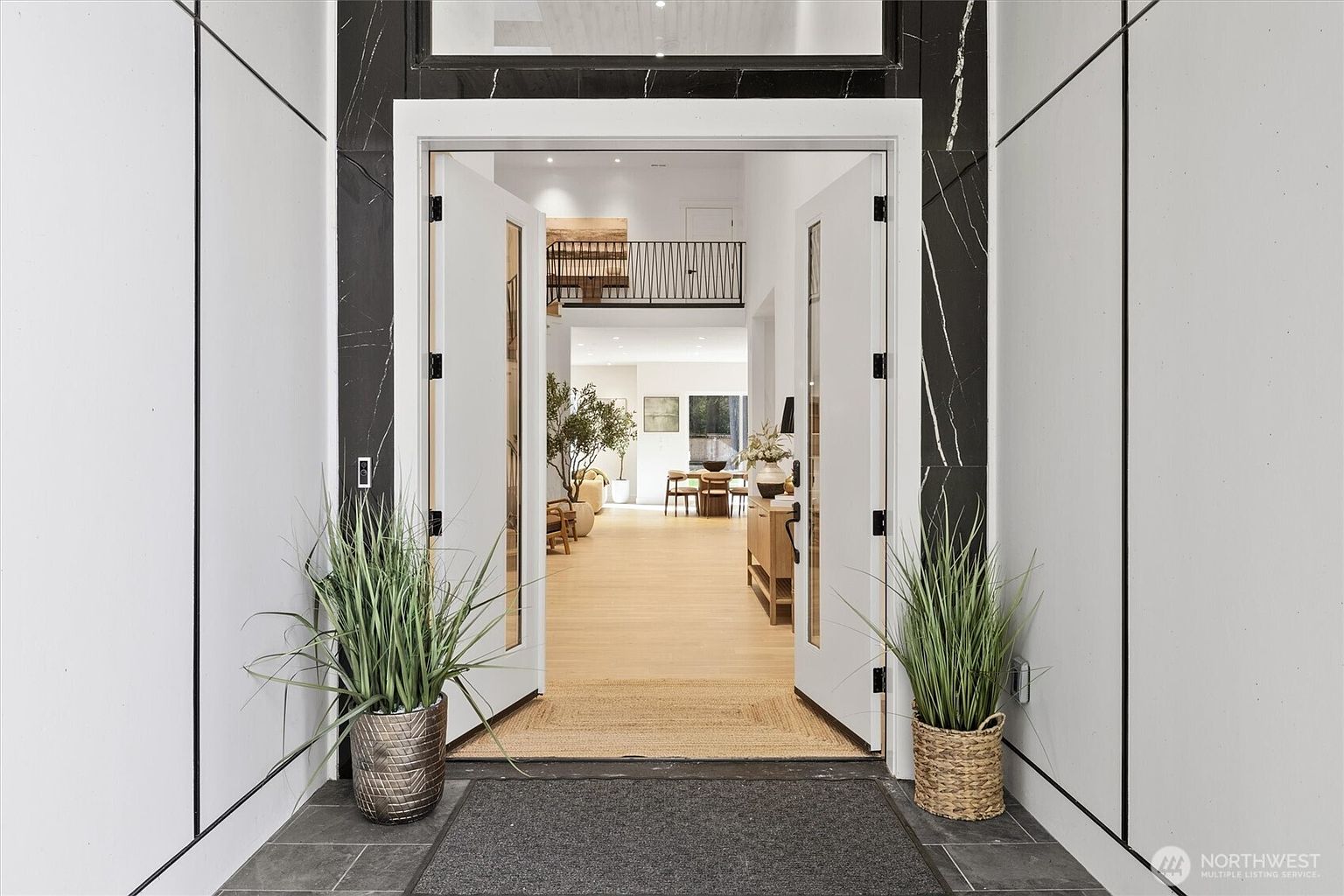 This is an interior shot of a home's entryway, showcasing an open floor plan with a view into the living and dining areas. The entrance features double doors flanked by potted plants, leading into a bright, modern space with hardwood floors and a staircase visible in the background. The perspective is from the entrance, inviting viewers into the home.