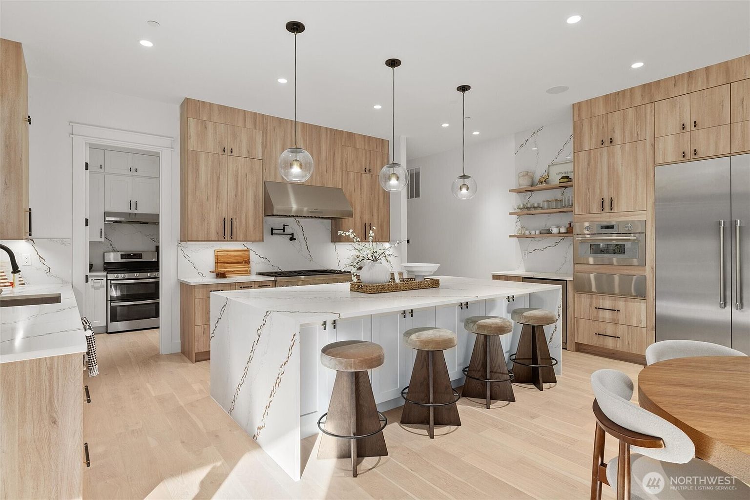 This is a bright and modern kitchen featuring light wood cabinetry, white countertops with marble veining, and stainless steel appliances. A large island with seating is the focal point, illuminated by pendant lights. The space is open and airy, creating a welcoming atmosphere.