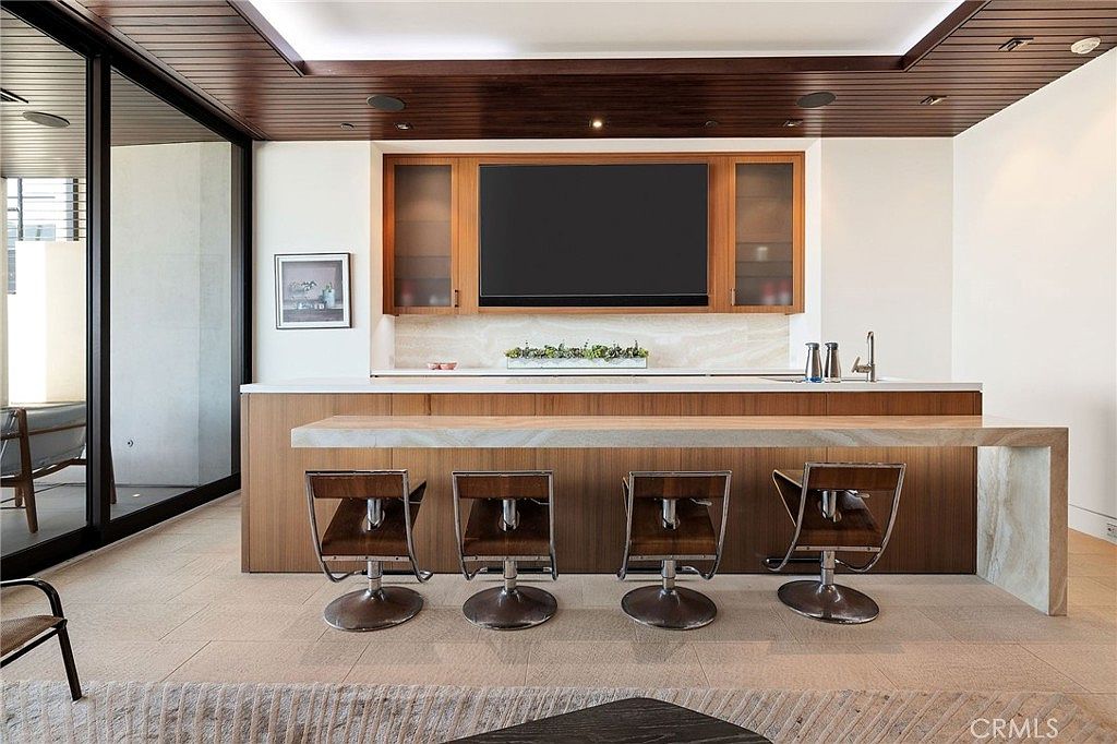 This modern wet bar or kitchen area features a sleek, wood-paneled bar with a long, light-toned stone countertop and four matching swivel stools. Behind the bar, a wall-mounted television is flanked by glass-front cabinetry, all set against a neutral stone backsplash. The space is defined by a dark wood-slatted ceiling and large glass sliding doors that open to an adjacent outdoor area, creating a seamless transition between indoor and outdoor living.
