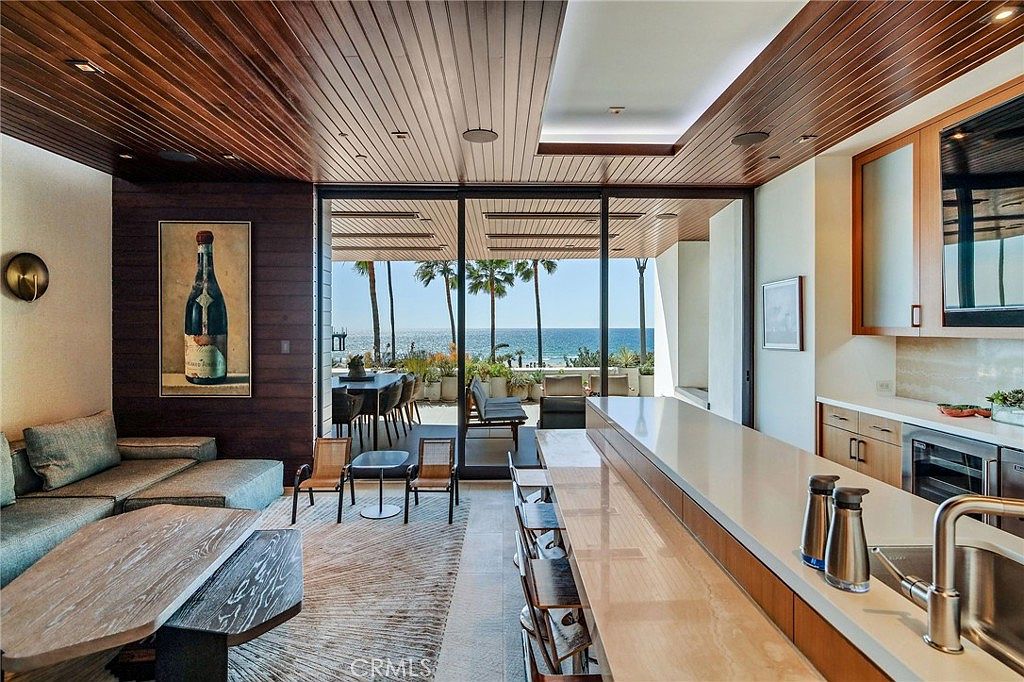 This open-concept living space seamlessly blends indoor and outdoor living, featuring a sleek kitchen island and a comfortable seating area. Large sliding glass doors open to a patio with ocean views, while the room is accented by a warm wood-paneled ceiling and a piece of art depicting a wine bottle. The perspective captures the flow from the kitchen through the living area to the coastal scenery beyond.