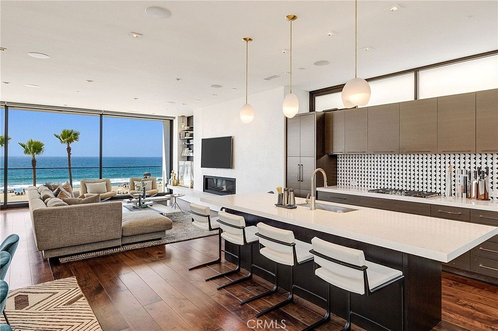 This open-concept living and kitchen area features a seamless transition to a stunning ocean view through expansive glass walls. The kitchen is equipped with dark cabinetry, a patterned tile backsplash, and a large central island with bar seating, while the adjacent living space offers a comfortable sofa and a fireplace. The room is bathed in natural light, highlighting the rich wood flooring and modern pendant lighting, creating a sophisticated and airy coastal atmosphere.