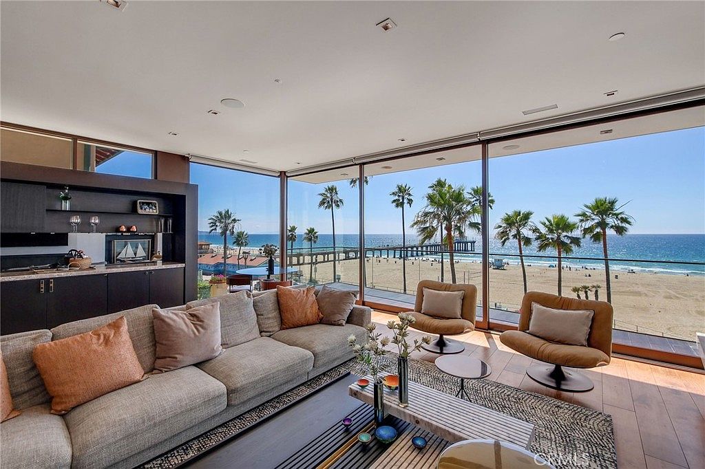 This sophisticated living room features a spacious seating area with a large gray sofa and two modern swivel chairs, all oriented toward a stunning panoramic view of the ocean and pier. The room is defined by expansive floor-to-ceiling glass walls that seamlessly blend the indoor living space with the beach scenery outside. A dark wood built-in bar area with open shelving adds a touch of luxury, while the warm wood flooring and textured area rug create a comfortable and inviting atmosphere.