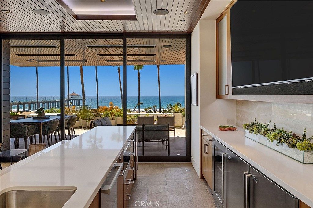This modern kitchen features a long island with a light-colored countertop and a built-in sink, complemented by a secondary counter area equipped with under-counter appliances and a television mounted above. Large sliding glass doors provide a seamless transition to an outdoor patio, framing a stunning, unobstructed view of the ocean and a nearby pier. The space is designed with a clean, contemporary aesthetic, utilizing neutral tones and natural light to create an airy, sophisticated atmosphere.