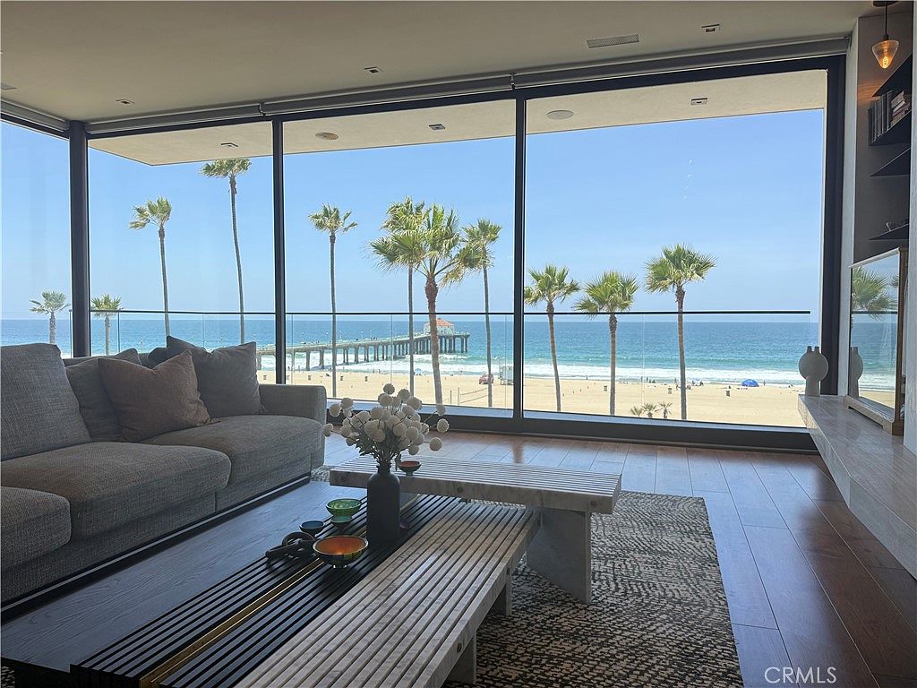 This modern living room features expansive floor-to-ceiling windows that provide a panoramic, unobstructed view of the ocean, beach, and a distant pier. The interior is furnished with a plush gray sofa, a low-profile wooden coffee table, and a textured area rug, creating a sophisticated and serene atmosphere. The space is bathed in natural light, emphasizing the seamless connection between the indoor living area and the coastal scenery outside.
