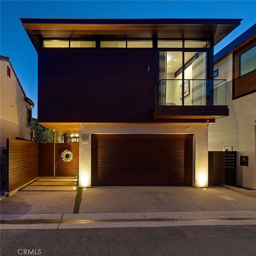 This modern two-story residence features a striking dark wood-paneled upper level contrasted against a light-colored stucco base. The front facade includes a wide garage door, a private pedestrian gate with a decorative wreath, and a glass-railed balcony on the second floor. Warm exterior lighting illuminates the driveway and entryway, creating an inviting and sophisticated curb appeal.