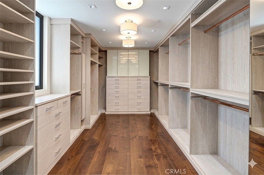 This spacious walk-in closet features custom light wood cabinetry with extensive shelving, hanging rods, and a central bank of drawers. The room is anchored by rich, dark wood flooring and illuminated by a tiered, modern light fixture. The symmetrical layout and clean lines create a highly organized and luxurious storage environment.
