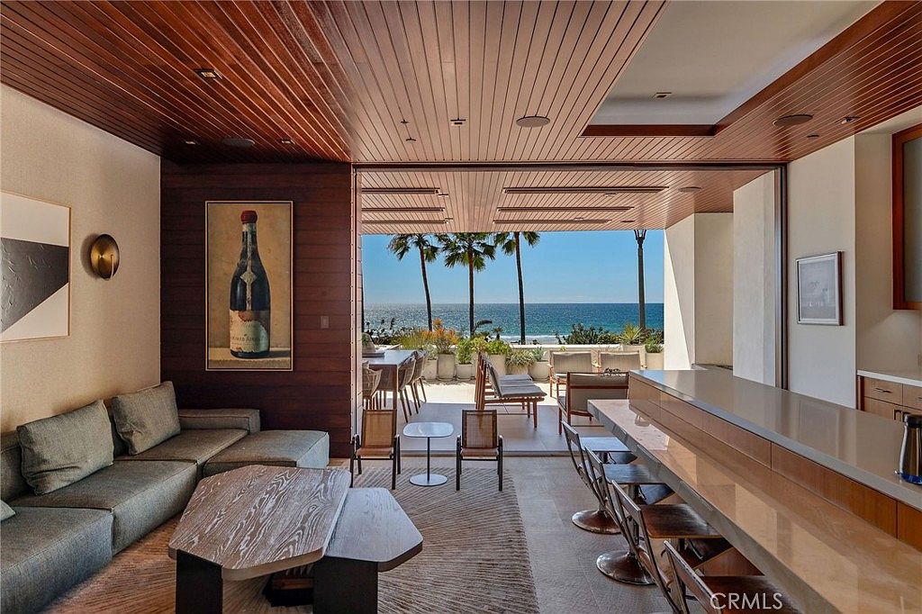 This open-concept living space seamlessly transitions to an outdoor patio, framing a stunning, unobstructed view of the ocean and palm trees. The interior features a comfortable gray sofa, a unique geometric coffee table, and a long bar counter with stools, all set beneath a warm wood-paneled ceiling. The design emphasizes indoor-outdoor living, with large glass openings that invite natural light and coastal breezes into the room.