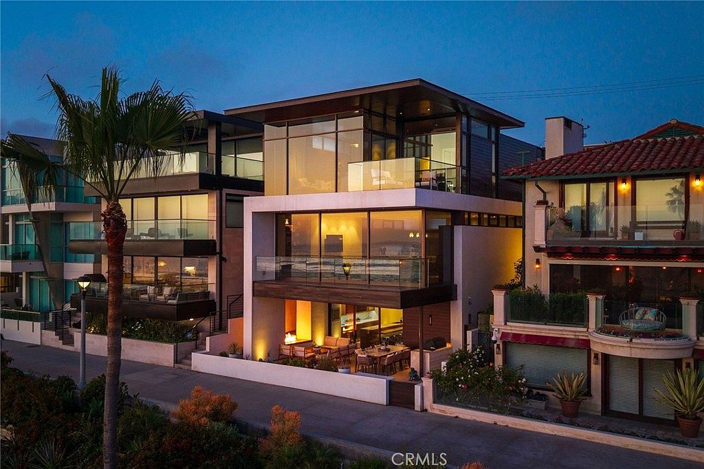 This striking contemporary beachfront residence features a multi-level design with extensive glass walls that offer panoramic ocean views. The home includes multiple levels of outdoor living space, including a ground-level patio with a fire feature and dining area, as well as upper-level balconies. The architectural style is modern and minimalist, emphasizing clean lines and an open, airy connection to the coastal environment.