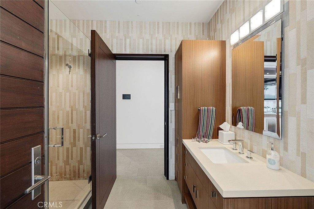 This modern bathroom features a sleek vanity with a light-toned countertop, a rectangular undermount sink, and a tall wood-toned storage cabinet. To the left, a glass-enclosed shower is visible, while a large rectangular mirror hangs above the vanity, reflecting the textured wall tiles. The perspective is from the doorway, looking into the bathroom space, which is illuminated by a multi-bulb light fixture above the mirror.