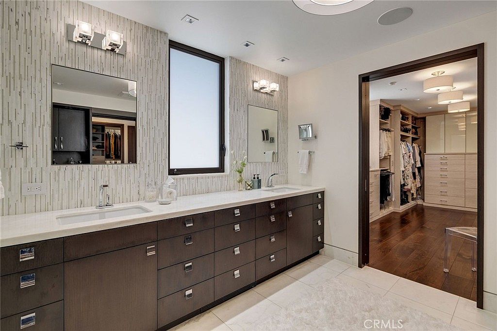 This primary bathroom features a long, dark-toned vanity with dual sinks and a light-colored countertop, set against a textured, vertical-patterned wall. A large window sits between the two mirrors, providing natural light, while an open doorway leads directly into a spacious, well-organized walk-in closet with dark wood flooring. The overall aesthetic is modern and sophisticated, with clean lines and a neutral color palette.