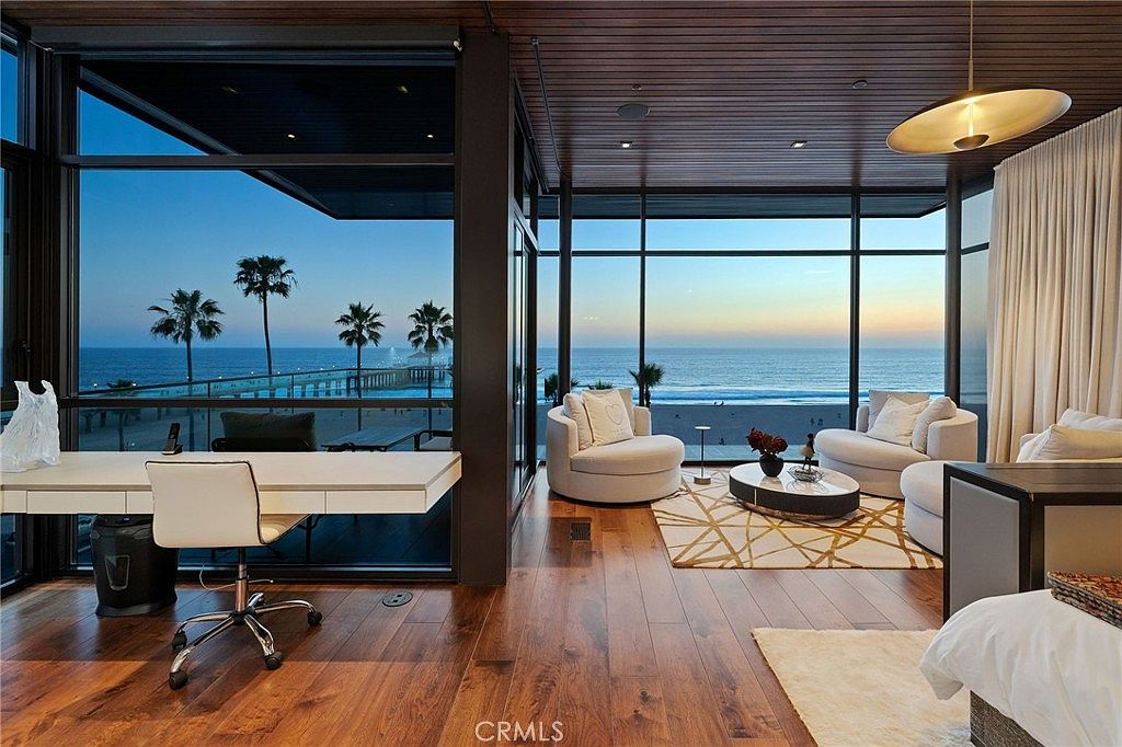 This sophisticated multi-purpose room features a seamless blend of a home office and a lounge area, defined by expansive floor-to-ceiling windows that offer panoramic ocean and pier views. The space is anchored by rich wood flooring and a warm, wood-paneled ceiling, creating a luxurious and serene atmosphere. A sleek white desk sits on the left, while a cozy seating area with two rounded armchairs and a circular coffee table occupies the right, all bathed in the soft, golden light of dusk.