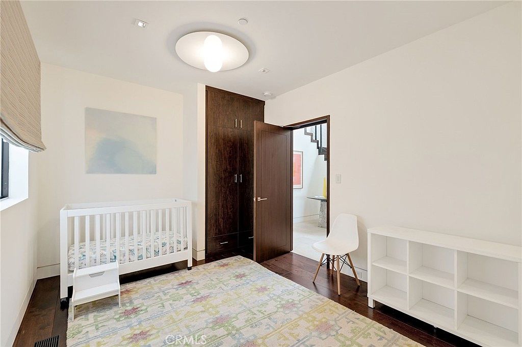This bright, modern nursery features dark wood flooring and a patterned area rug, centered around a white crib. A tall, dark wood built-in cabinet provides storage, while a white open-shelf unit sits against the wall. The room is illuminated by a circular ceiling fixture and natural light from a window on the left, creating a clean and minimalist aesthetic.