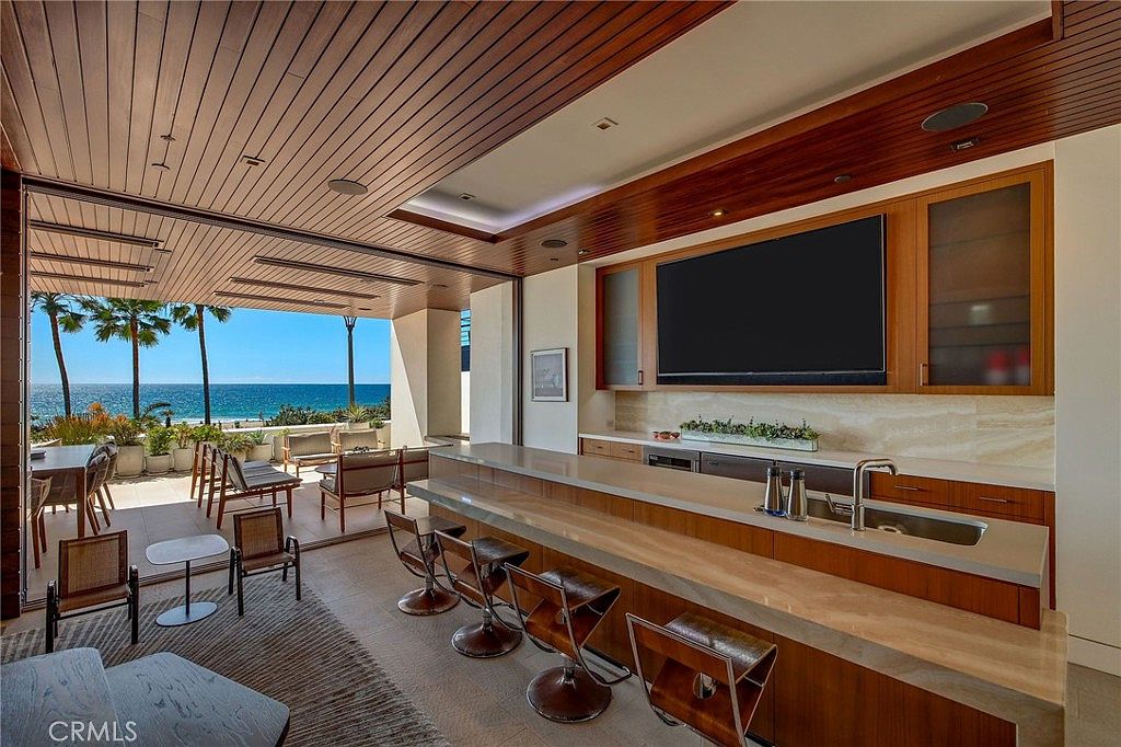 This indoor-outdoor living space features a sleek bar area with a long stone countertop and modern stools, seamlessly transitioning to a covered patio through a wide opening. The room is accented by a warm wood-paneled ceiling and a large wall-mounted television, while the adjacent outdoor area offers a direct, unobstructed view of the ocean and palm trees. The overall aesthetic is luxurious and contemporary, designed to maximize the coastal scenery.