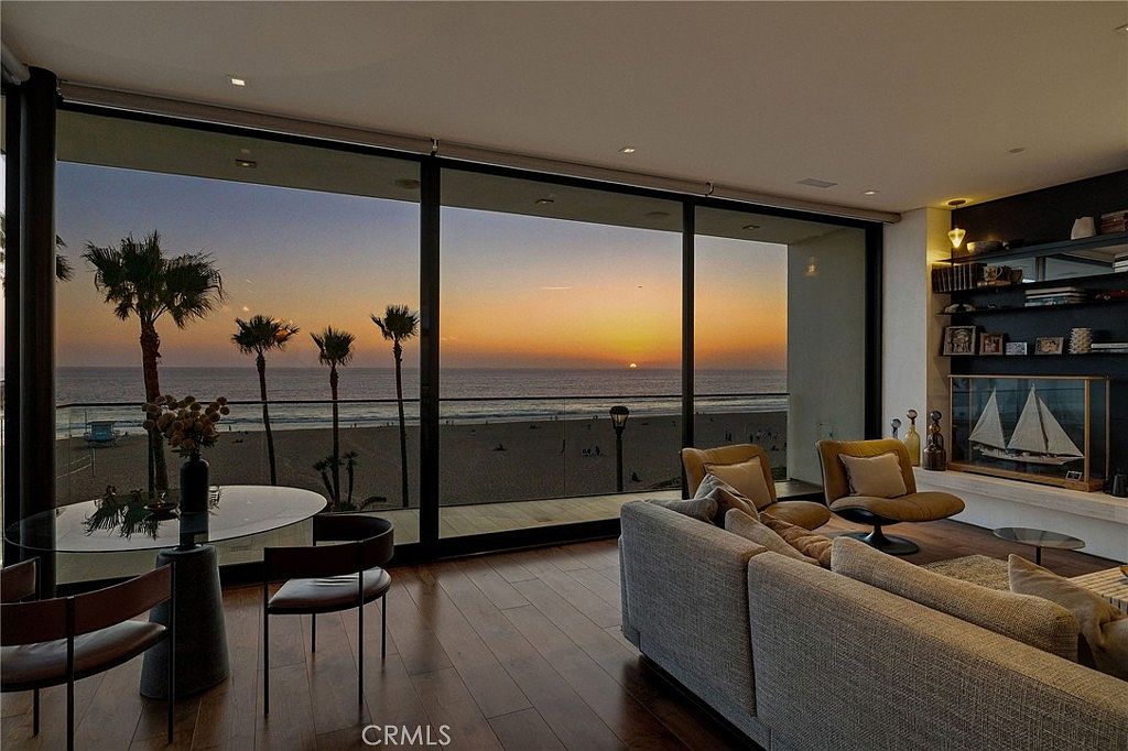 This sophisticated living space features an open-concept layout with dark wood flooring and a neutral color palette. The room is defined by expansive floor-to-ceiling glass walls that provide a panoramic view of the beach and sunset, seamlessly blending indoor and outdoor living. A comfortable grey sectional sofa and modern armchairs are arranged to face the ocean, while a built-in shelving unit with a decorative model ship adds a refined touch to the interior.