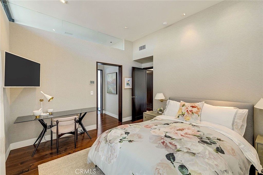 This bright and modern bedroom features a queen-sized bed with a floral-patterned duvet and a neutral upholstered headboard. To the left, a sleek glass-topped desk with a minimalist chair creates a dedicated workspace, complemented by a wall-mounted television. The room is finished with dark wood flooring, a light-colored area rug, and a doorway leading to an adjacent hallway, all under a high, vaulted ceiling.